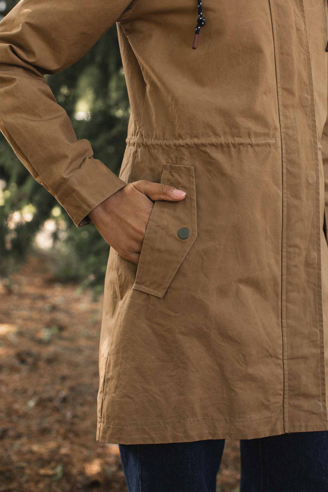 A tan Briar Jacket / Dark Khaki Waxed Canvas hooded parka with a drawstring waist and a buttoned flap pocket, shown with a hand inside the pocket.
