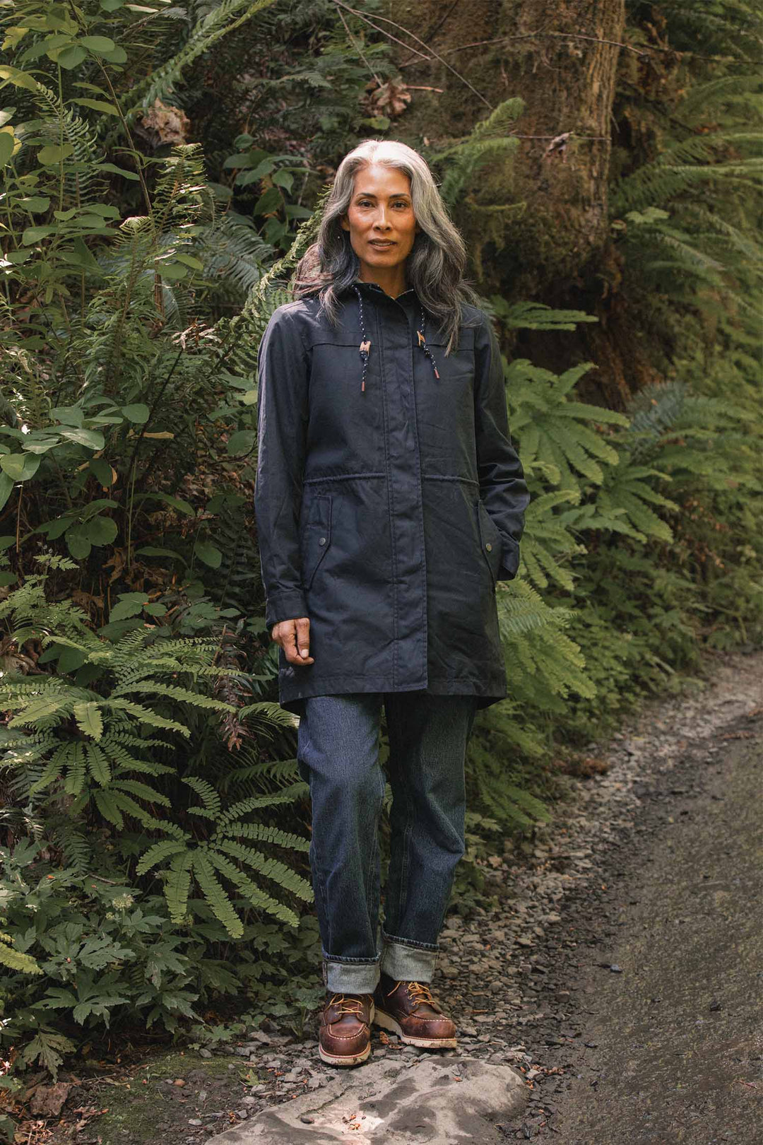 A person wearing a navy, hooded Bridge & Burn Briar Jacket / Navy Waxed Canvas with a drawstring waist, jeans, and leather boots standing on a forest path.