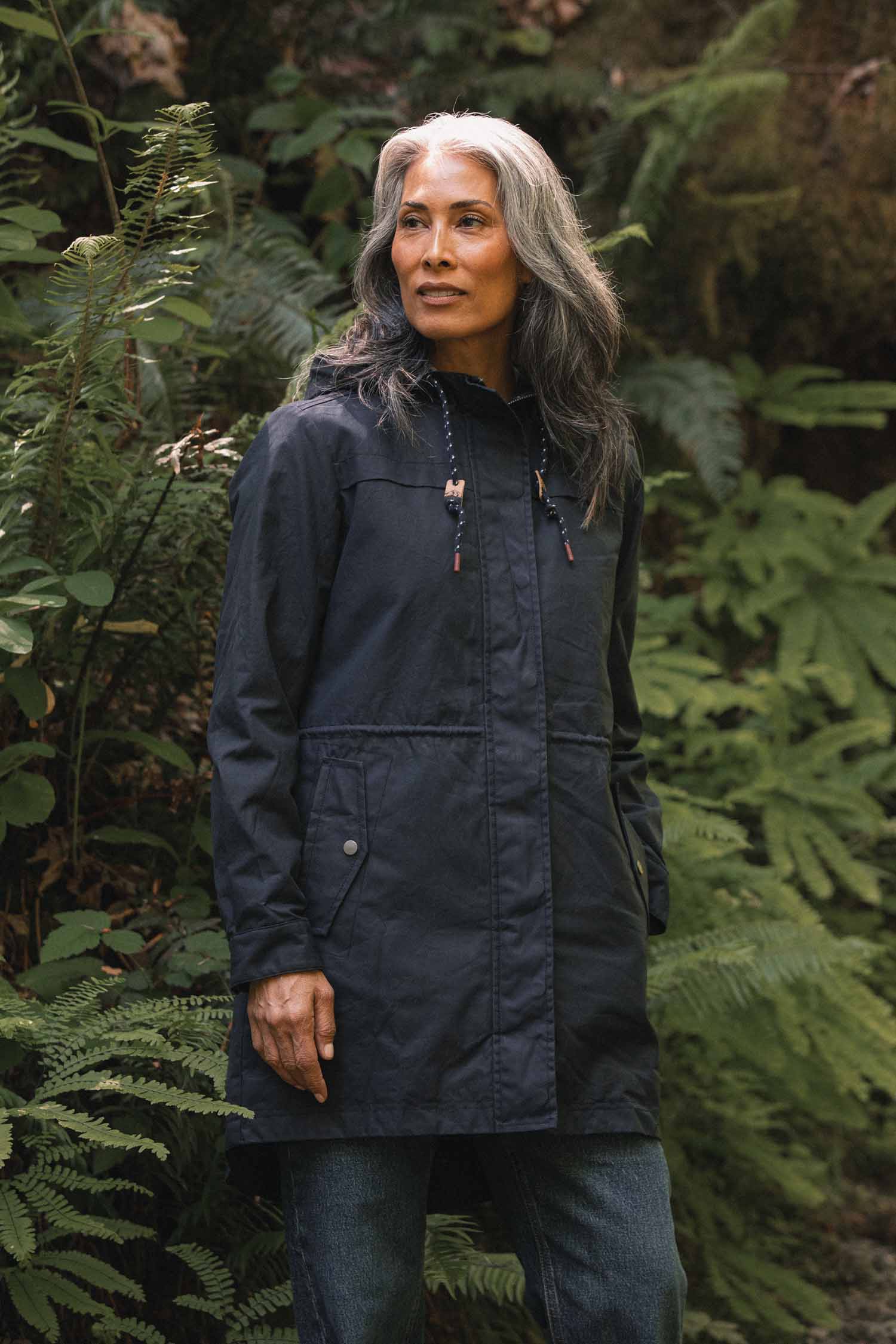 A woman wears a navy waxed canvas Briar Jacket, a rain-ready hooded parka with a drawstring waist, standing in a forest.
