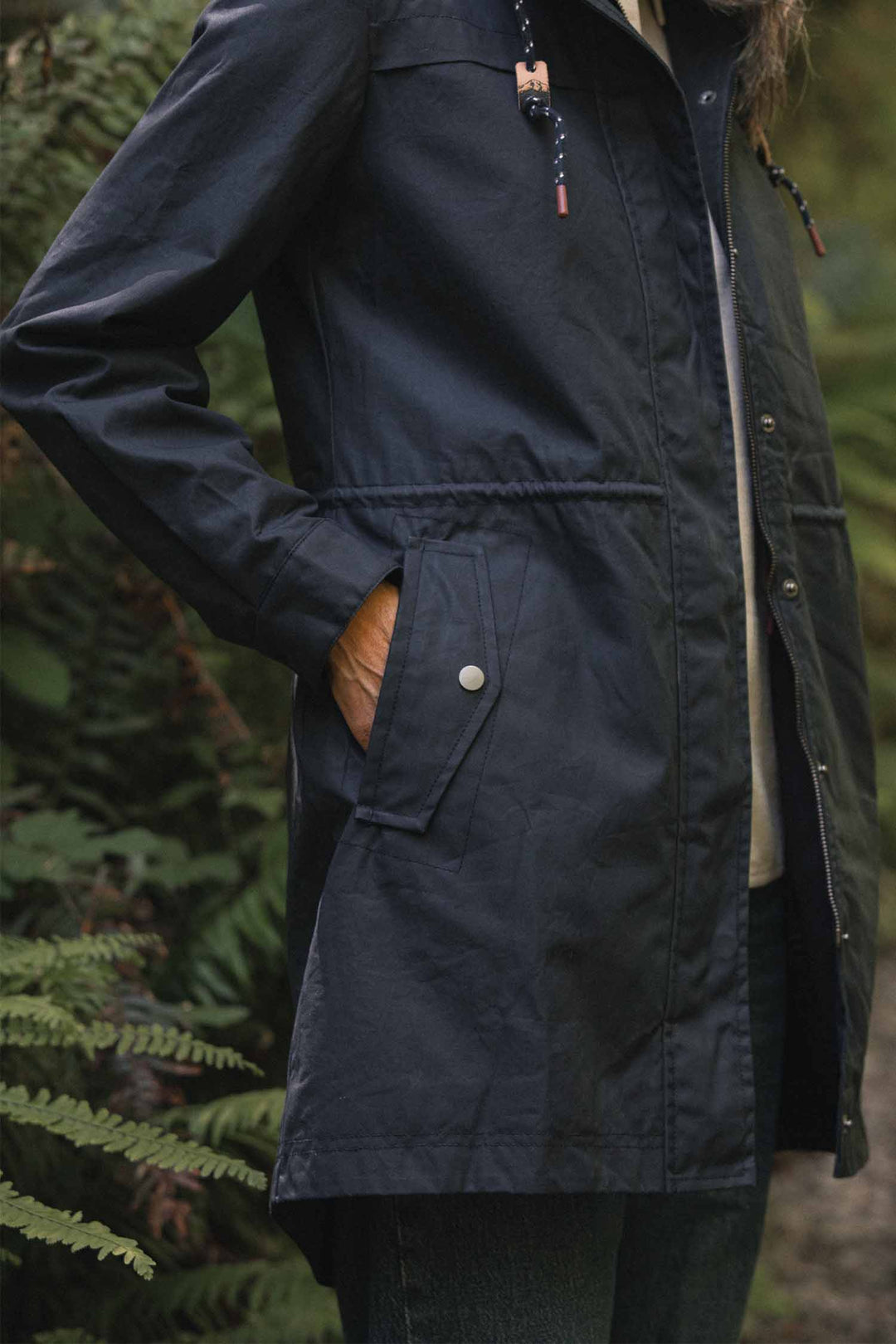 A person wearing a navy, rain-ready Bridge & Burn Briar Jacket / Navy Waxed Canvas hooded parka with a drawstring waist and a snap-button pocket.