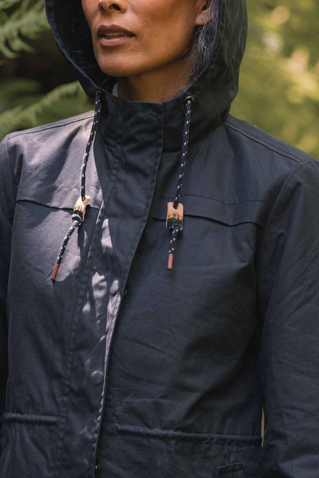 A person wearing a navy Bridge & Burn Briar Jacket / Navy Waxed Canvas hooded parka with rope drawstrings held by patterned wooden toggles.