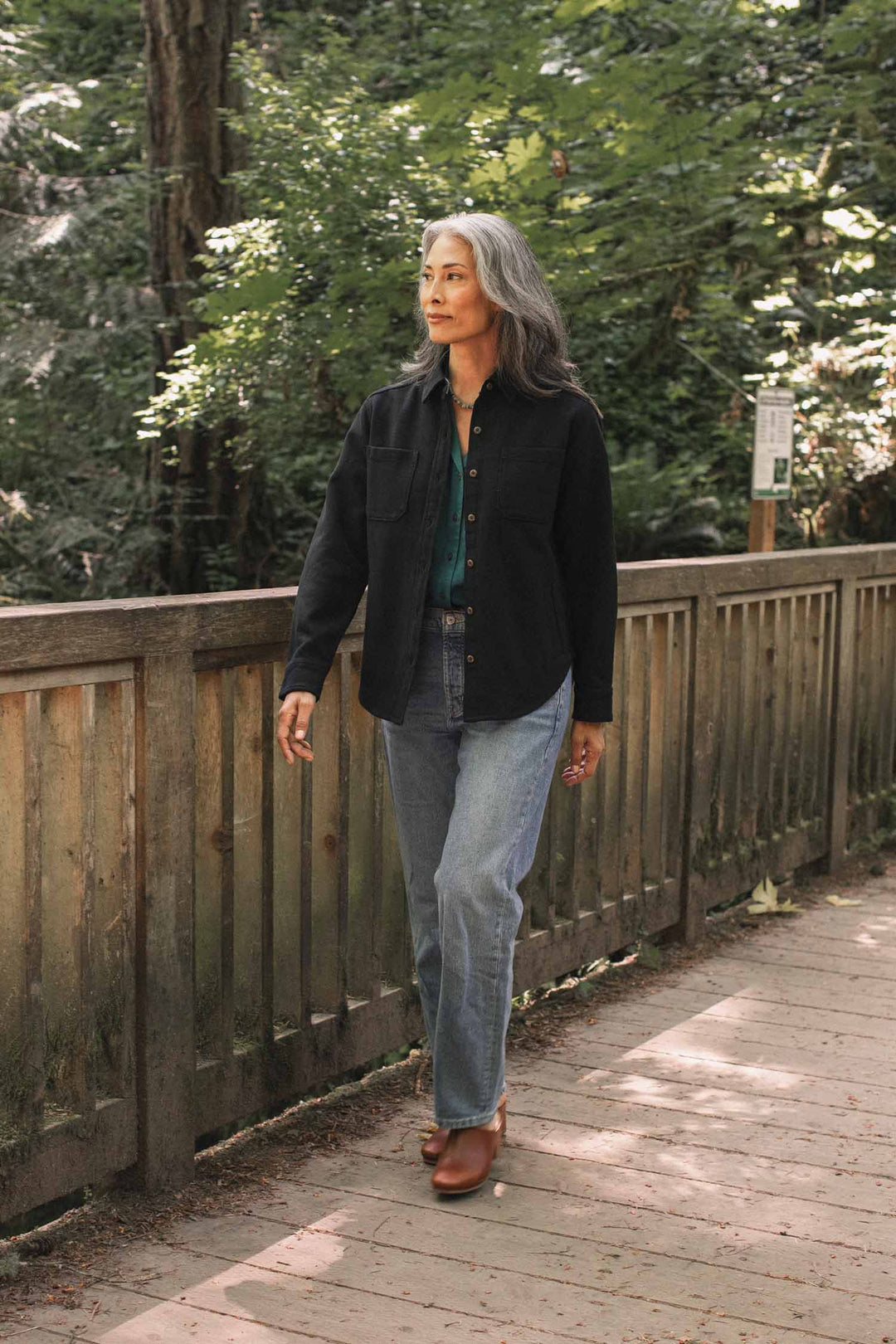 A woman with long gray hair walks along a wooden path through a sun-dappled forest, wearing the Bridge & Burn Dawson Overshirt in Ink Stone. Dressed in comfortable, sustainable clothing made from recycled cotton, she pairs her black jacket with a teal shirt, blue jeans, and brown shoes as sunlight filters through the green trees around her.