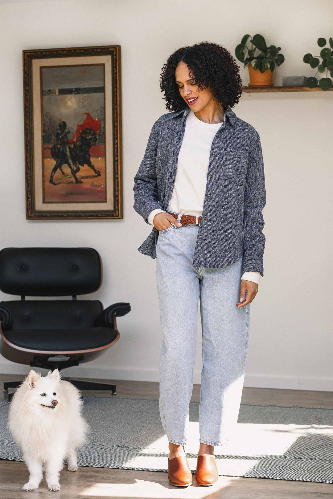 A woman stands indoors with a fluffy white dog, dressed in an effortlessly stylish look featuring the Bridge & Burn Dawson Overshirt in Ocean Marl—a chic and sustainable choice made from recycled cotton. She pairs it with a white shirt, jeans, and brown shoes. In the background, there’s a black chair, a painting on the wall, and a shelf adorned with potted plants.
