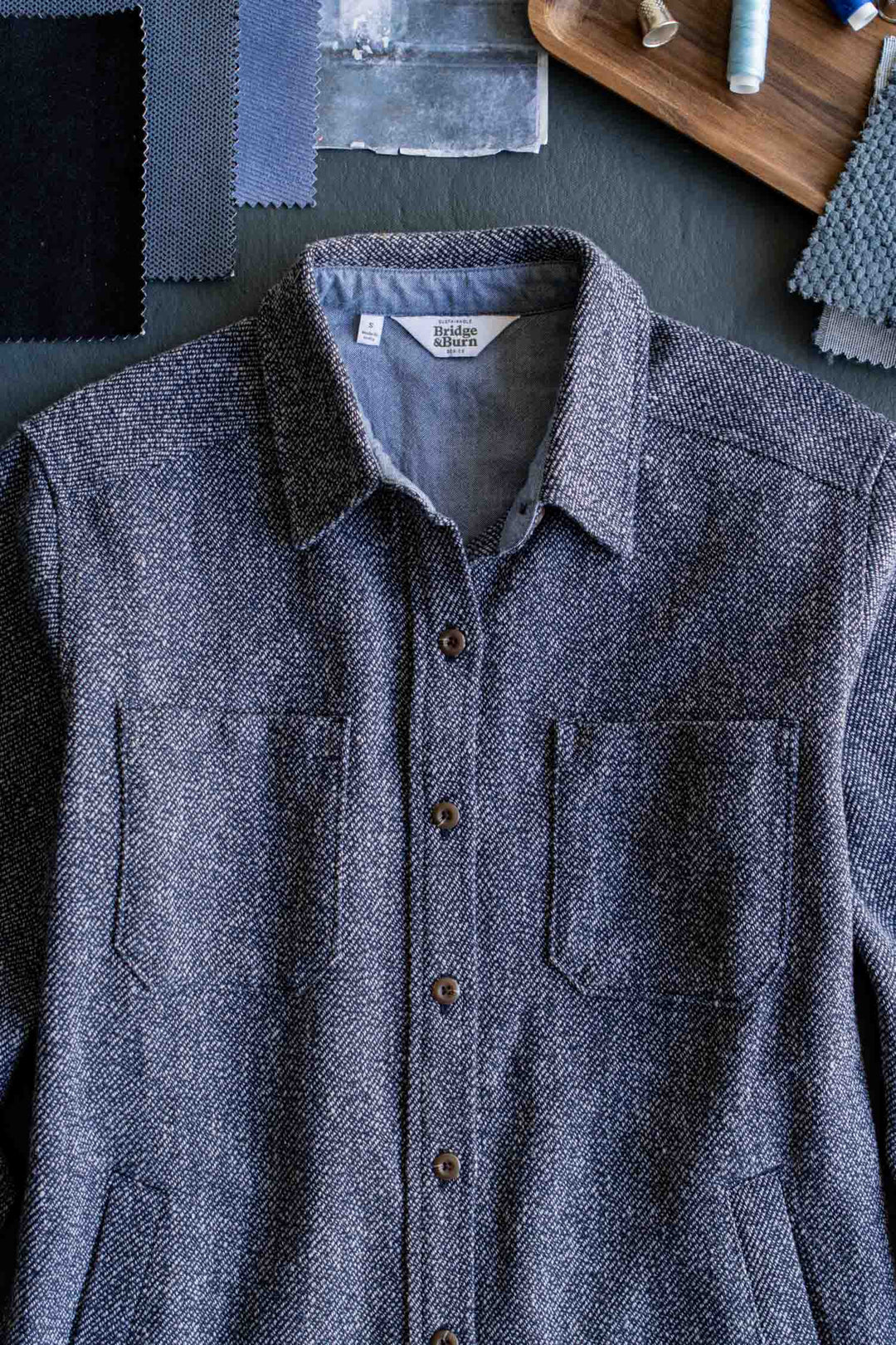 The Dawson Overshirt in Ocean Marl by Bridge & Burn, a textured gray button-up crafted from recycled cotton, is laid flat on a dark surface and surrounded by fabric swatches, sewing thread spools, and other sewing materials.