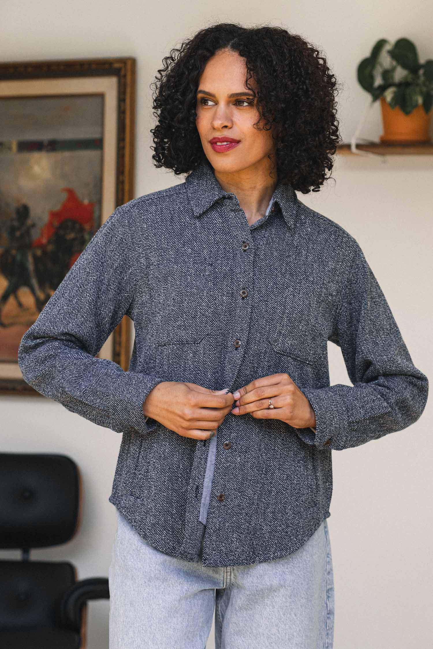 A person with curly hair wearing the Bridge & Burn Dawson Overshirt in Ocean Marl and light jeans stands indoors, tying the waist of their shirt. A painting and a potted plant in the background highlight their sustainable choice.