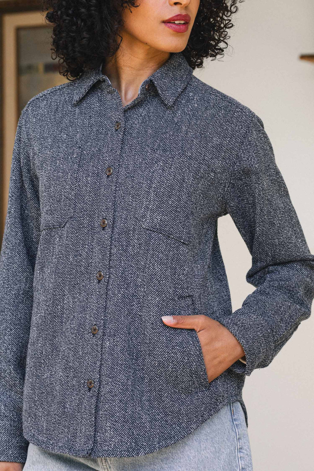A woman stands indoors with her hand in her pocket, wearing the Bridge & Burn Dawson Overshirt in Ocean Marl—a textured gray recycled shirt featuring two chest pockets. The photo subtly highlights this sustainable style, showing only her torso and part of her face.