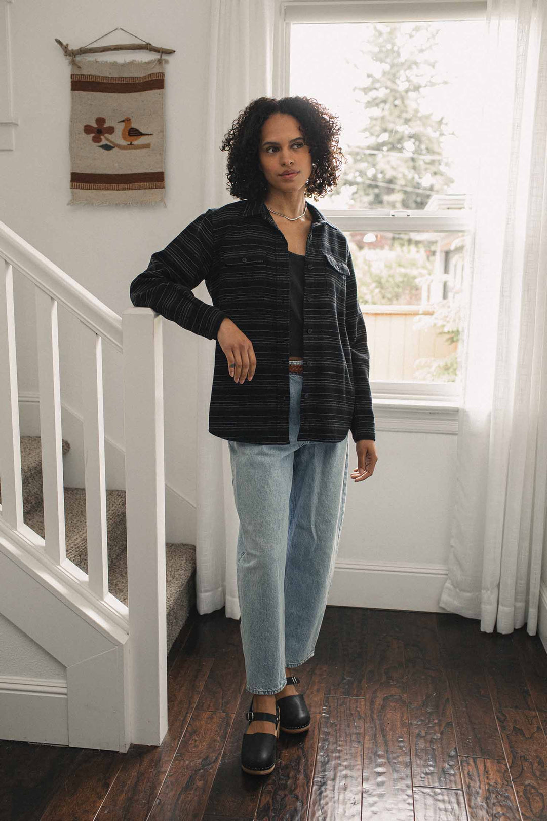A woman wearing a relaxed fit, horizontally striped Bridge & Burn Lenon Shirt / Raven Stripe layered over a black top and light-wash jeans.