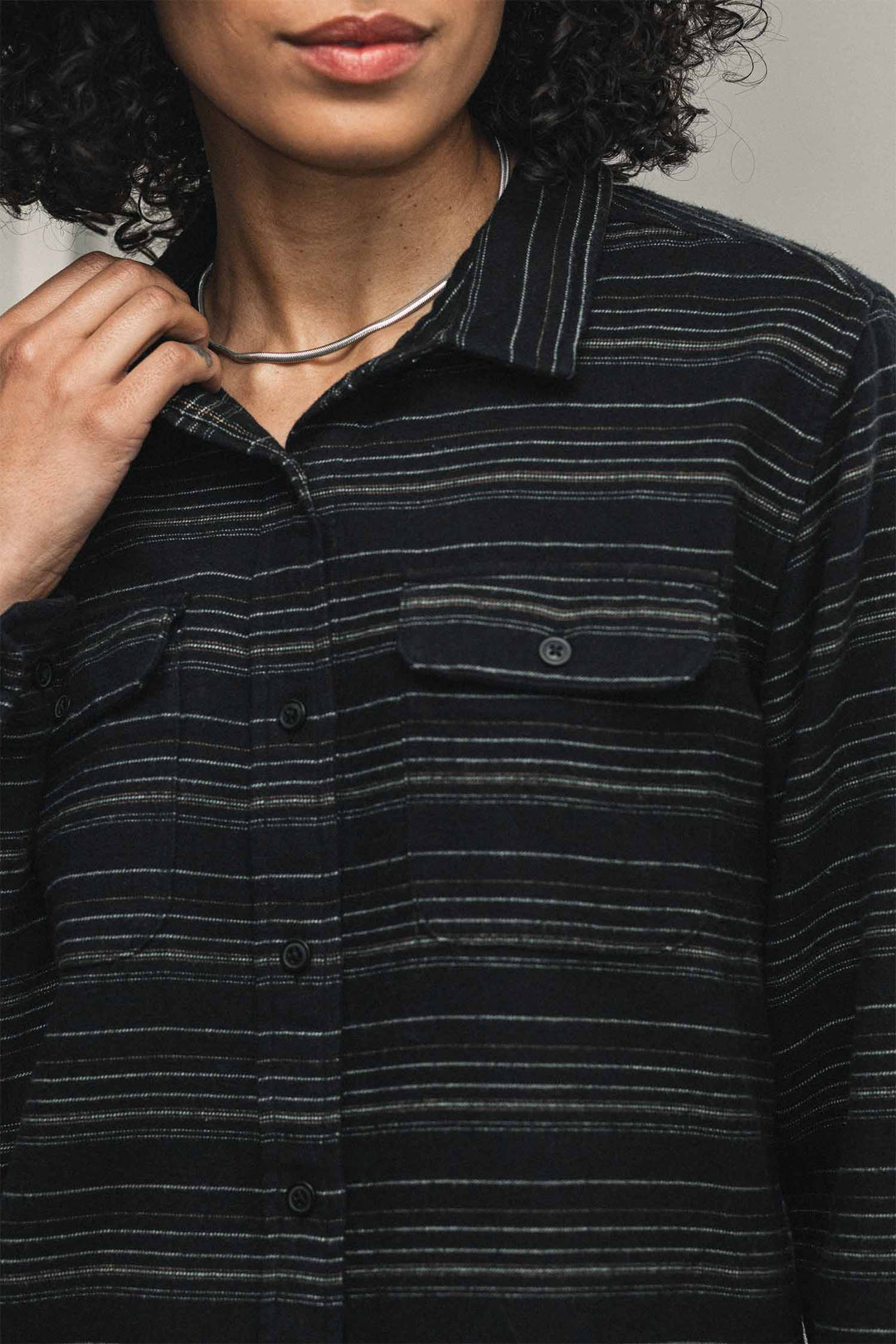 A person wears a Bridge & Burn Lenon Shirt / Raven Stripe, a relaxed fit black long-sleeved button-down over-shirt featuring thin, horizontal white stripes and two button-flap chest pockets.