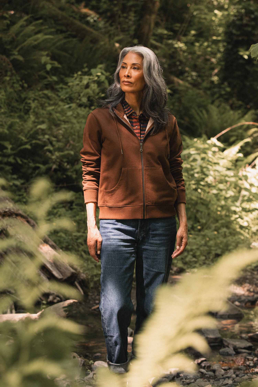 A person with long gray hair stands outdoors in a forest, wearing the Bridge & Burn Mason Hoodie in Cumin—a sustainable brown hoodie made from recycled cotton—paired with jeans, surrounded by greenery and sunlight filtering through the trees.