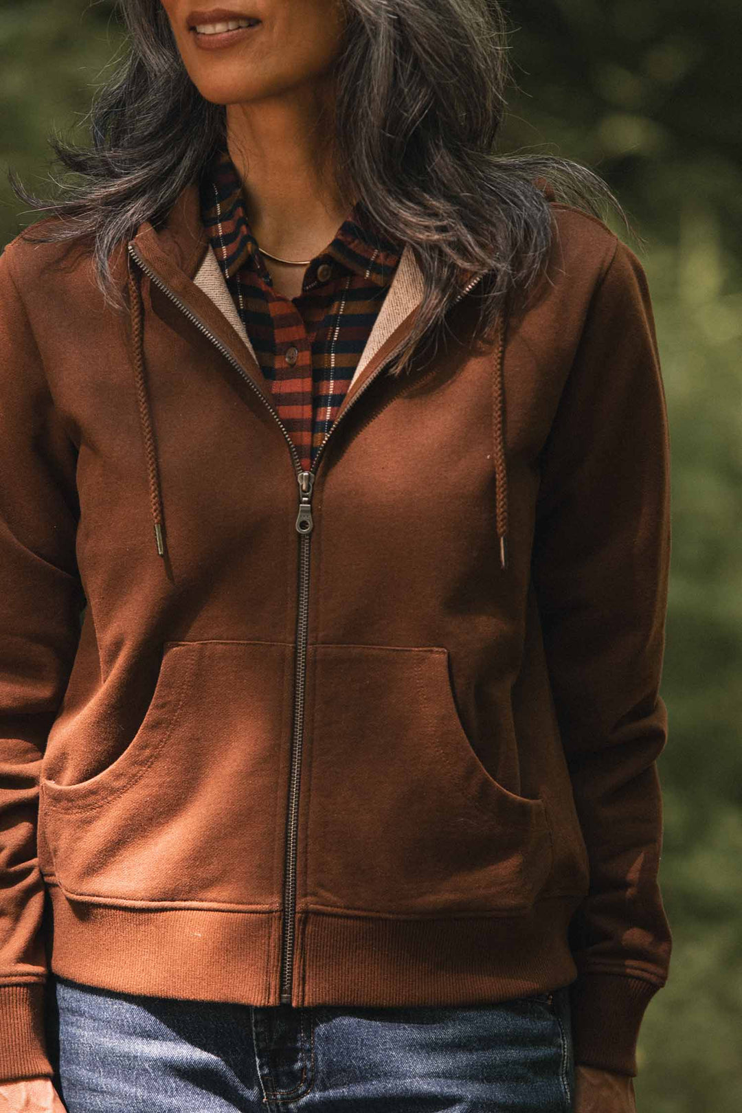 Wearing the Bridge & Burn Mason Hoodie in Cumin—crafted from recycled cotton for sustainability—a person stands outdoors over a plaid shirt, with only the lower half of their face visible.