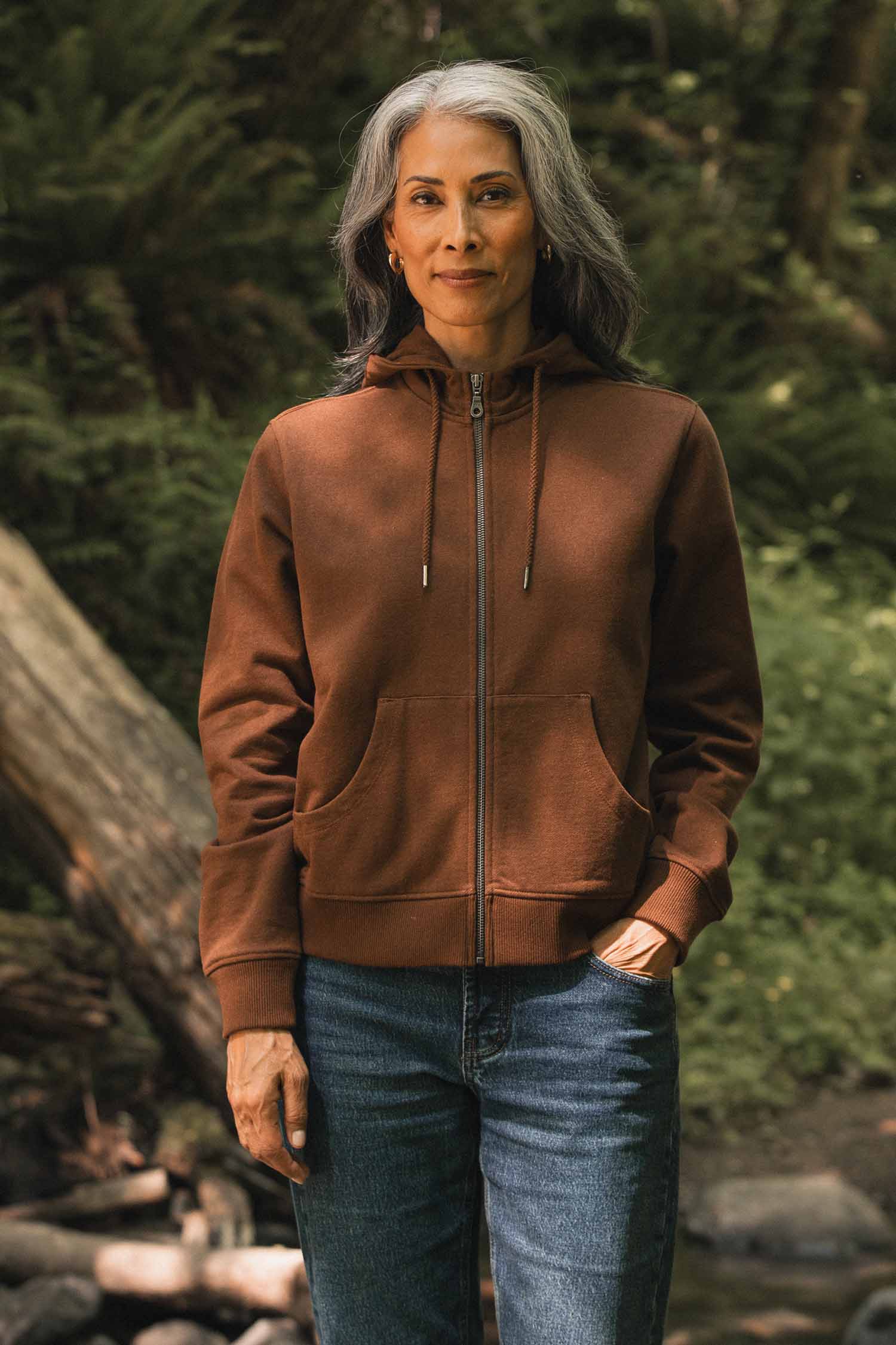Wearing the Bridge & Burn Mason Hoodie in Cumin, a person with long gray hair stands outdoors in jeans, surrounded by lush green forest scenery.