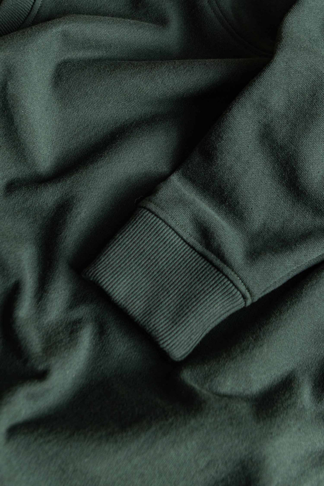 Close-up of the Bridge & Burn Mason Hoodie in Juniper, a dark green recycled French terry sweatshirt, highlighting the ribbed cuff of the sleeve resting on soft, slightly wrinkled fabric.