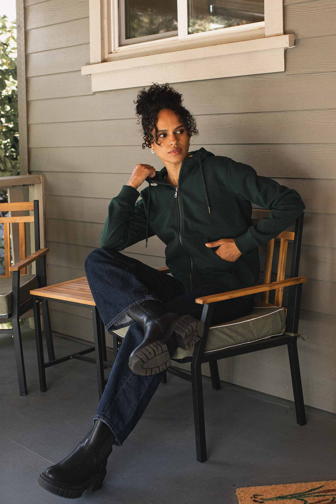Wearing the Bridge & Burn Mason Hoodie in Juniper—a dark green, sustainable hoodie crafted from recycled cotton—a person with curly hair sits on a patio chair on a porch. Dressed in dark jeans and black boots, they look to the side with one hand resting thoughtfully on their chin.