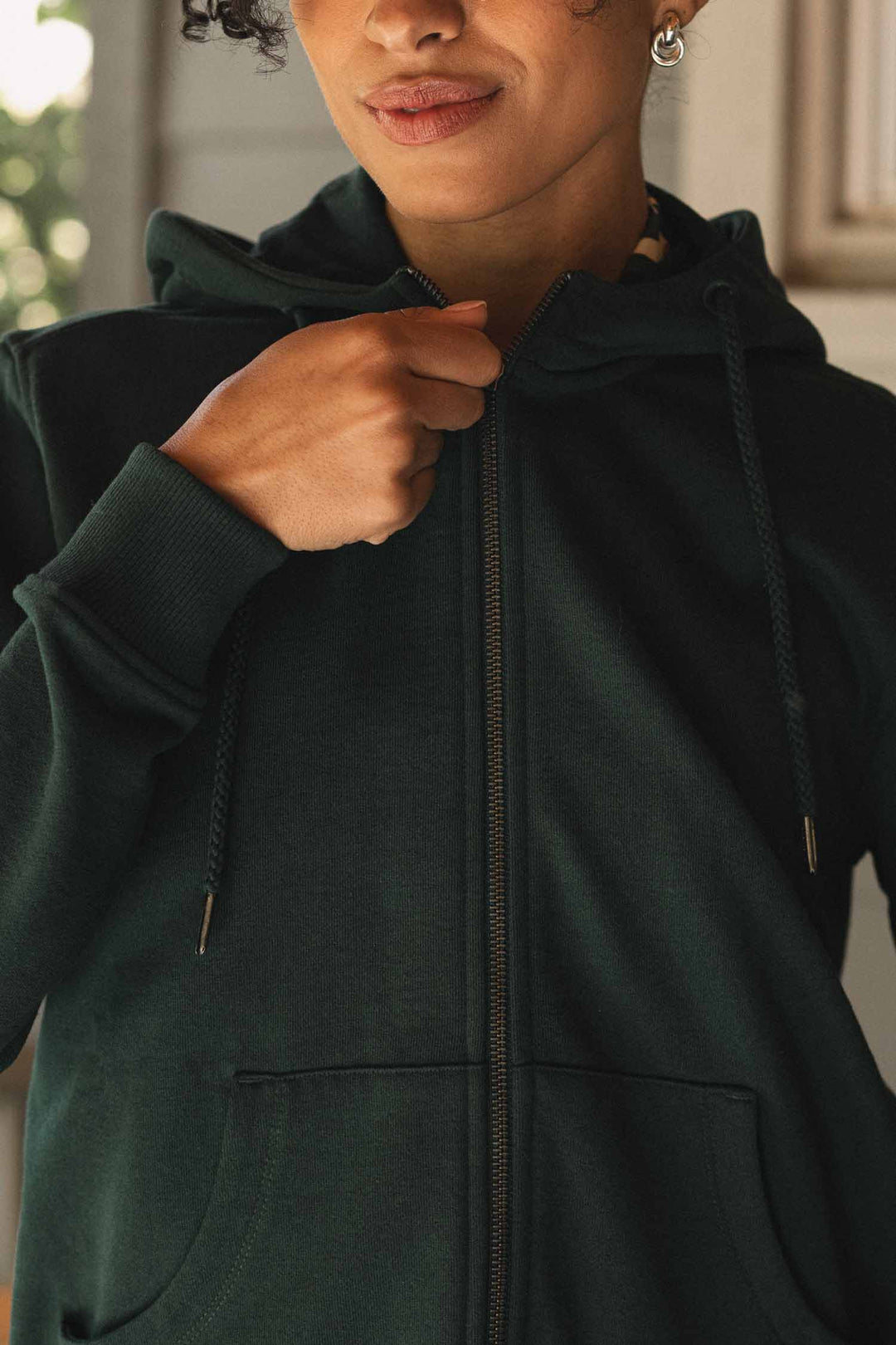 A person wearing the Bridge & Burn Mason Hoodie in Juniper holds the zipper near their chest. The image highlights the upper body, showing part of their face, hand, and torso.