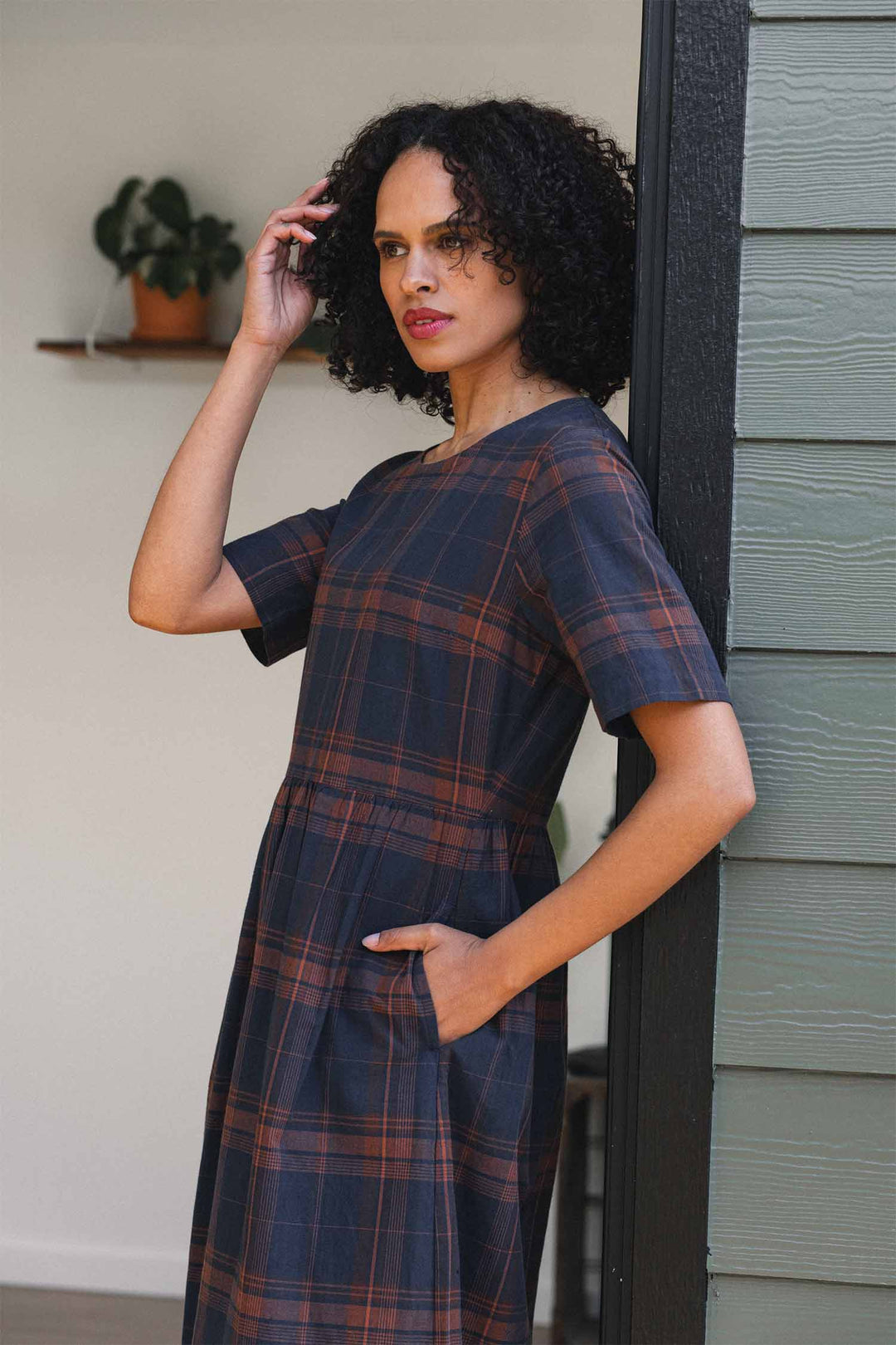 A woman with curly hair stands by a doorway, one hand touching her hair and the other in her pocket, wearing the Bridge & Burn Perry Dress in Cinder Plaid—an organic cotton dress featuring short sleeves, pockets, and a gathered skirt. A potted plant sits in the background.