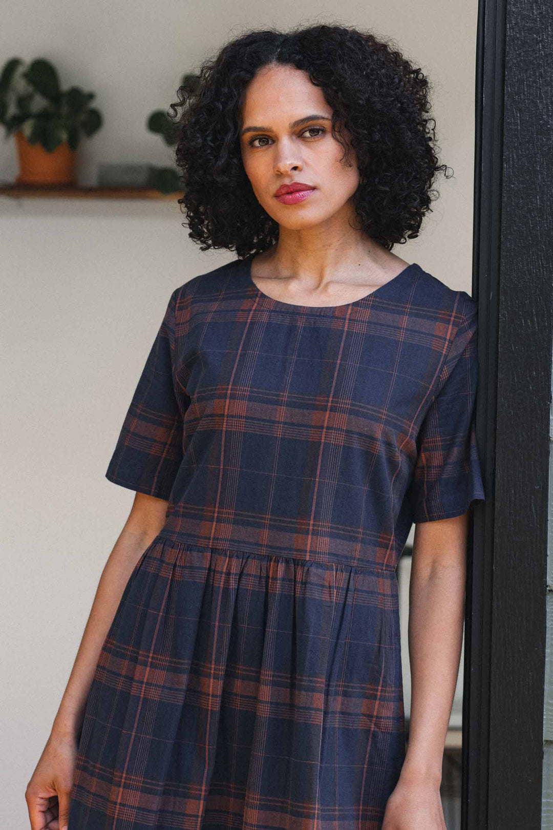A woman with curly hair wears the Bridge & Burn Perry Dress in Cinder Plaid and leans against a black doorframe. A potted plant rests on a shelf in the softly blurred background.