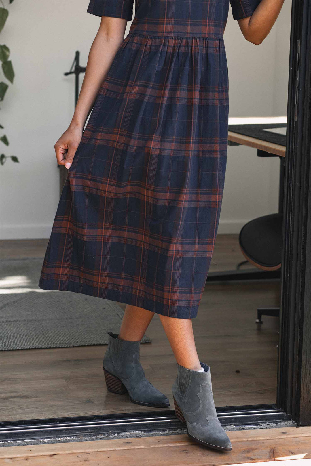 A person stands partially inside, holding the skirt of the Bridge & Burn Perry Dress in Cinder Plaid—an organic cotton dress with a gathered skirt—paired with gray ankle boots; one arm is bent and not fully visible.
