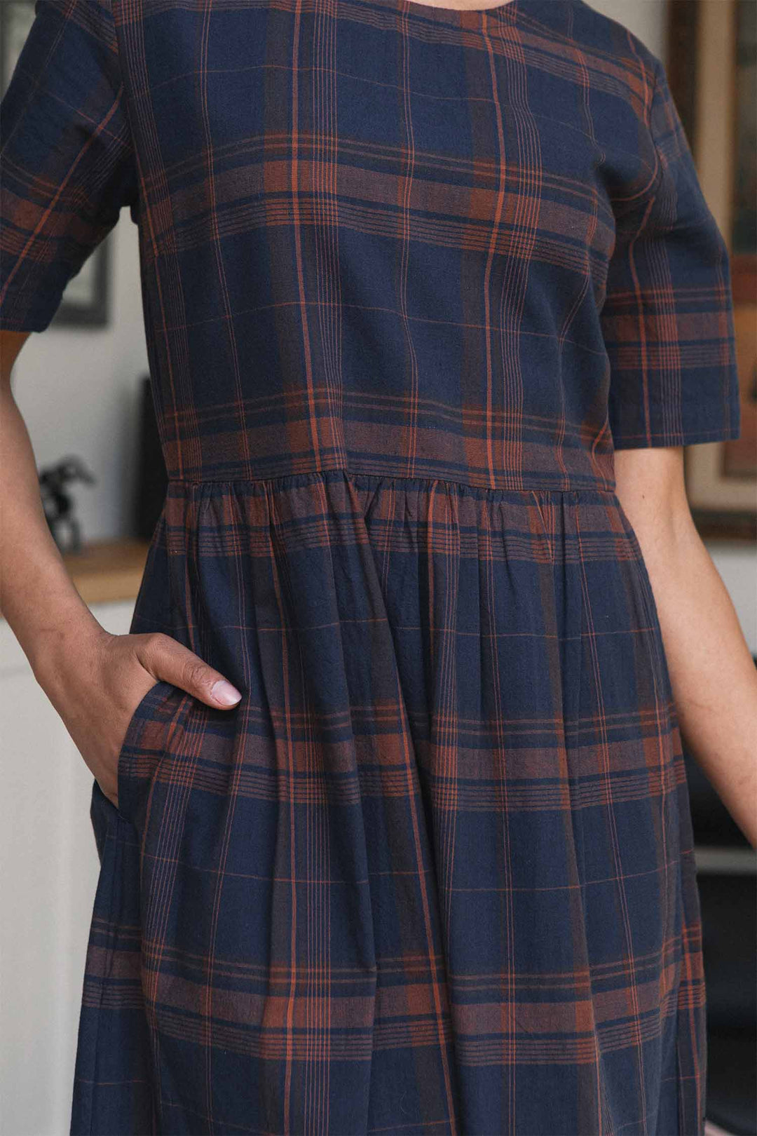 A person stands indoors with one hand in a side pocket, wearing the Bridge & Burn Perry Dress in Cinder Plaid. This organic cotton midi-length dress features a navy blue and rust plaid pattern, short sleeves, and a gathered skirt that adds subtle volume. The photo is cropped at the neck and knees.