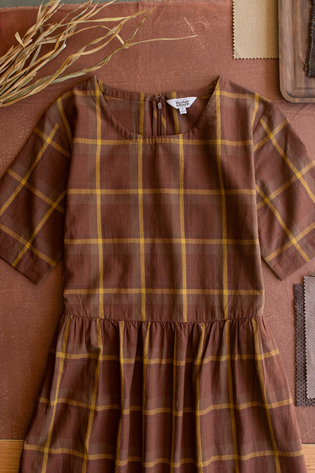 The Perry Dress in Harvest Plaid by Bridge & Burn is displayed flat on a brown surface, surrounded by dried grasses and fabric swatches. This midi-length dress features a charming silhouette with short sleeves, yellow stripes within its brown plaid pattern, and a gathered skirt at the waist.