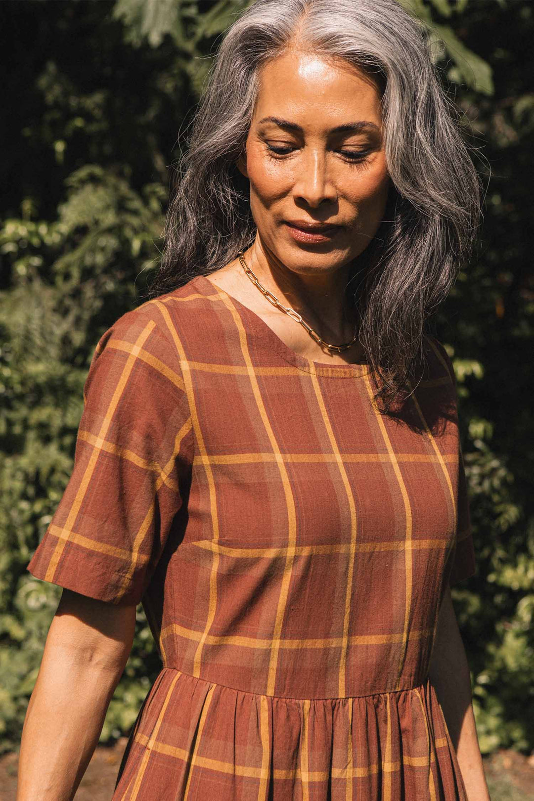 A woman with long gray hair stands outdoors amid greenery, looking down with a slight smile, wearing the Bridge & Burn Perry Dress in Harvest Plaid.