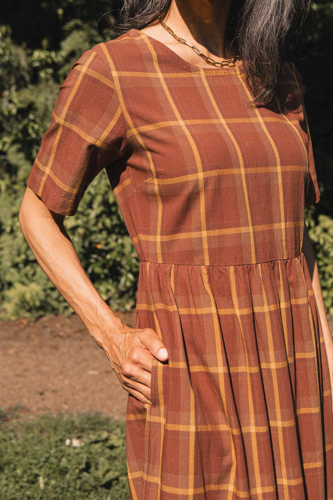 A woman wearing the Bridge & Burn Perry Dress in Harvest Plaid—an organic cotton dress featuring short sleeves and a gathered skirt—stands outdoors with one hand in her pocket, surrounded by green foliage and sunlight.