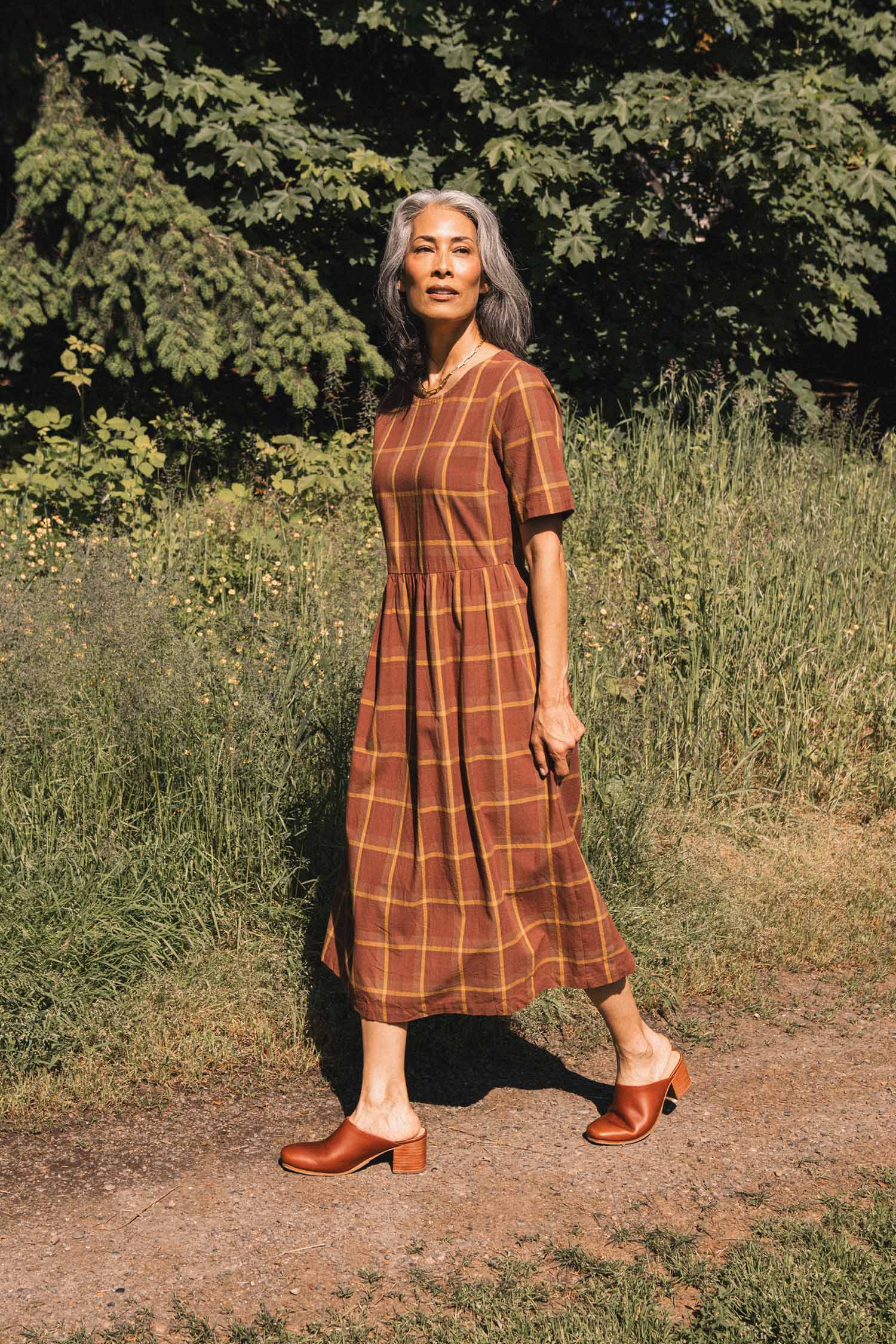 A woman with gray hair walks along a path surrounded by grass, wearing the Bridge & Burn Perry Dress in Harvest Plaid, featuring a gathered skirt, paired with brown shoes. Trees and greenery fill the background.
