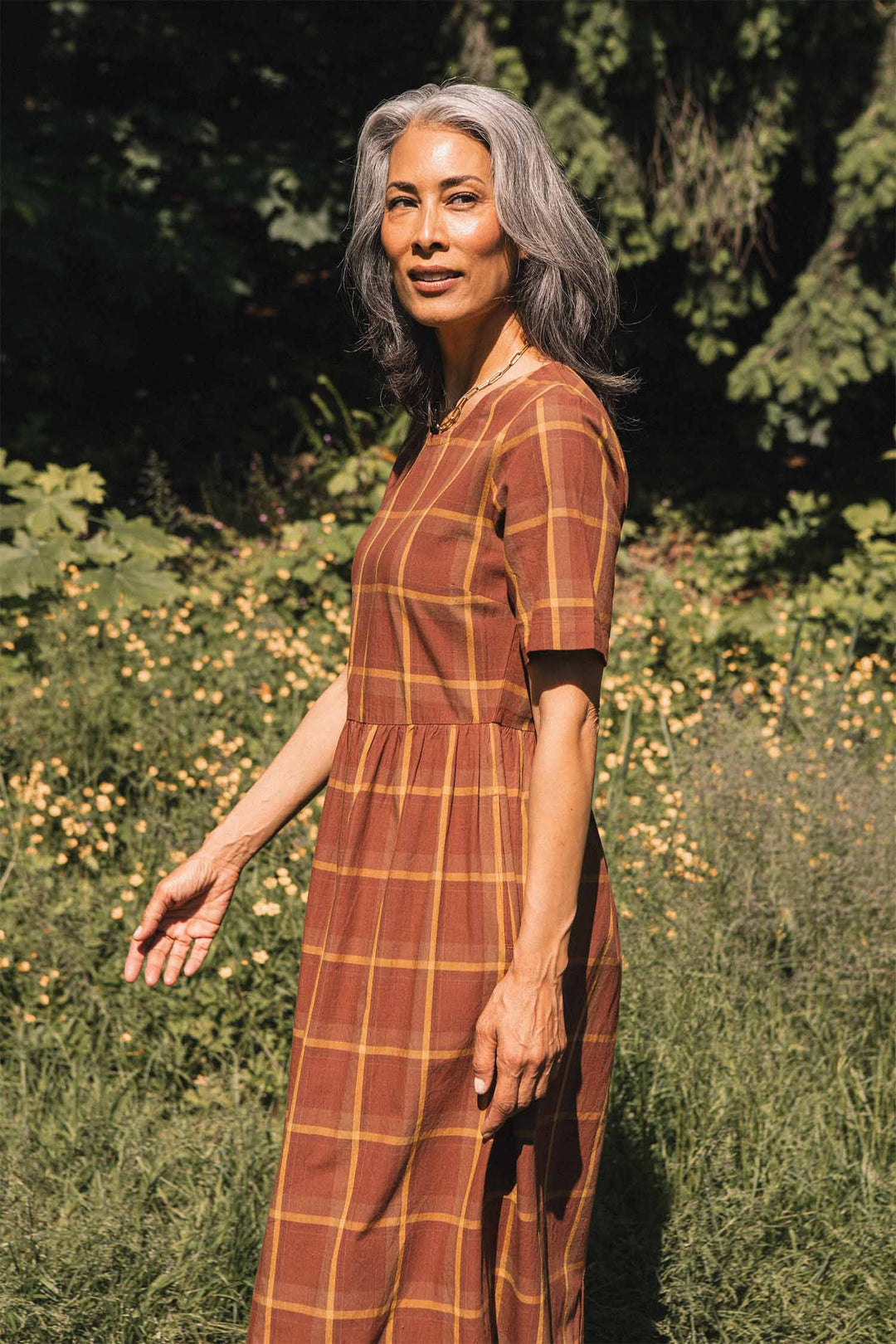 A woman with gray hair stands outdoors in a sunlit garden surrounded by green plants and yellow flowers, wearing the Bridge & Burn Perry Dress in Harvest Plaid. She looks at the camera with a relaxed expression, the organic cotton dress featuring a gathered skirt.