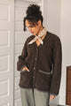 A woman stands indoors near a white paneled door, wearing the Bridge & Burn Whit Cardigan in Dark Olive—a cozy V-neck cardigan with gray patch pockets and textured knit details—paired with olive pants and a cream patterned scarf tied around her neck.