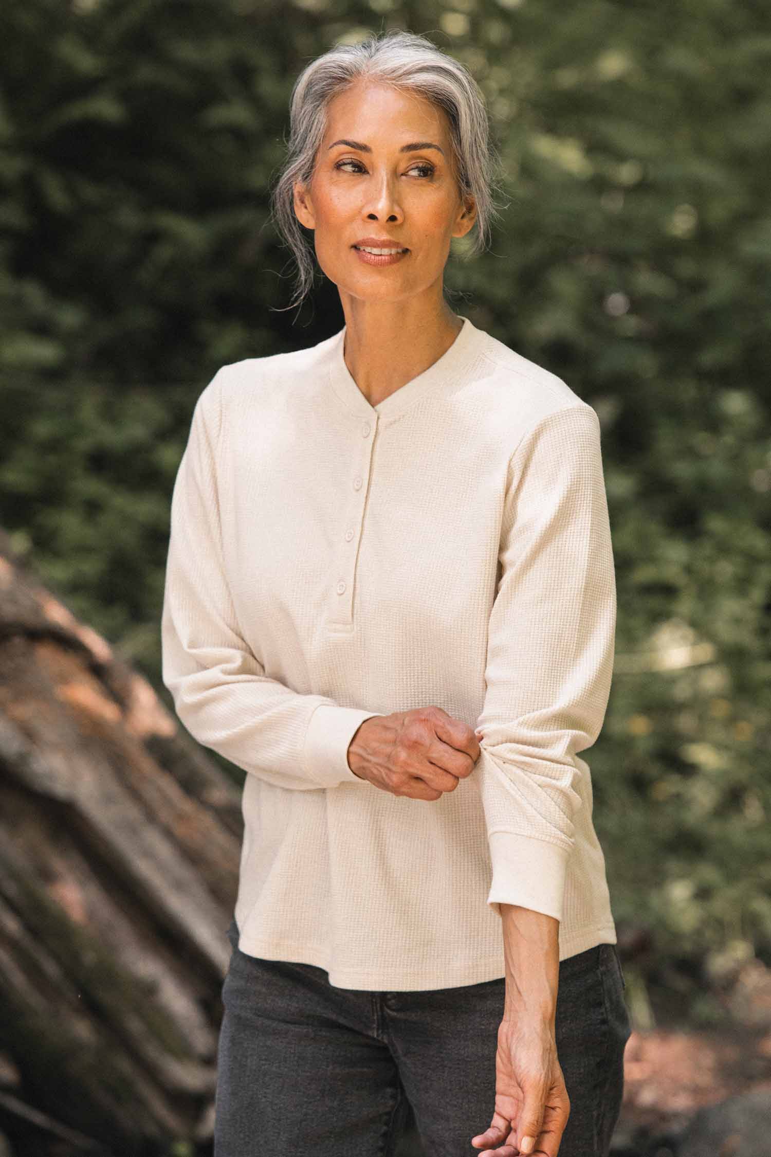 A woman with gray hair in a ponytail stands outdoors amidst green foliage and a fallen log, gently pulling up her sleeve. She is wearing the Bridge & Burn Women’s Recycled Waffle Henley in Ivory with dark pants.