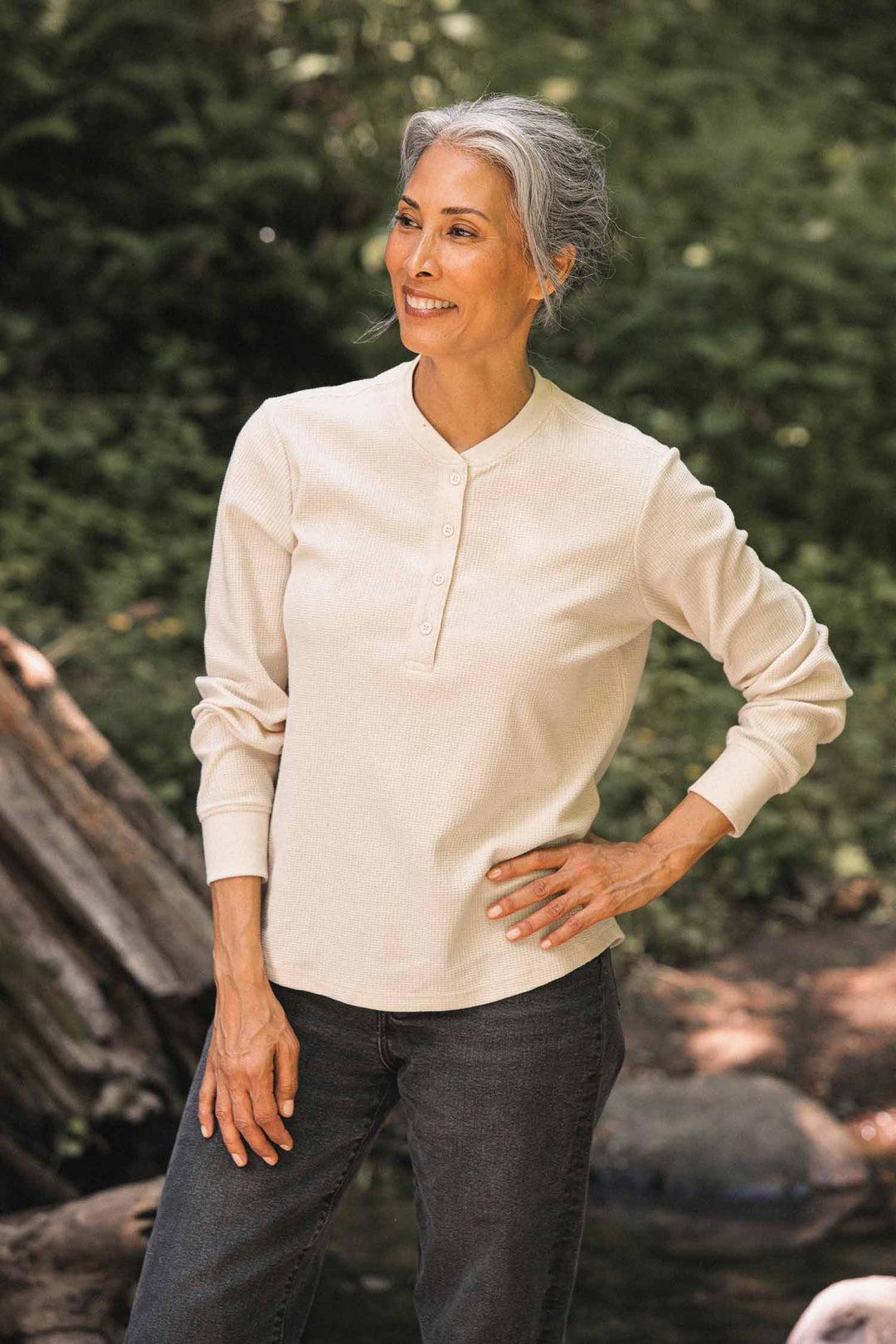 A woman with gray hair stands outdoors, smiling, wearing the Bridge & Burn Women’s Recycled Waffle Henley in Ivory and dark jeans, with trees and rocks in the background.