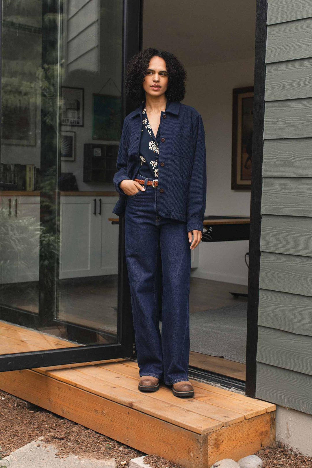 A person with curly hair stands in a doorway, wearing the Bridge & Burn Women’s Studio Chore Coat in Indigo over a floral shirt, paired with navy pants and brown shoes. The background reveals part of a modern house interior.