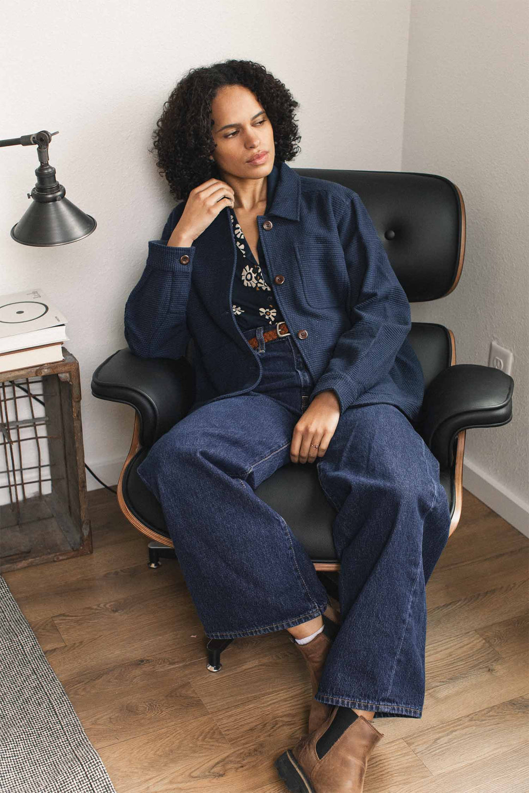 A person with curly hair sits on a black lounge chair, wearing the Bridge & Burn Women’s Studio Chore Coat in Indigo, paired with wide-leg jeans, brown boots, and a floral shirt. Nearby are a lamp, books, and a side table in a cozy, modern room.