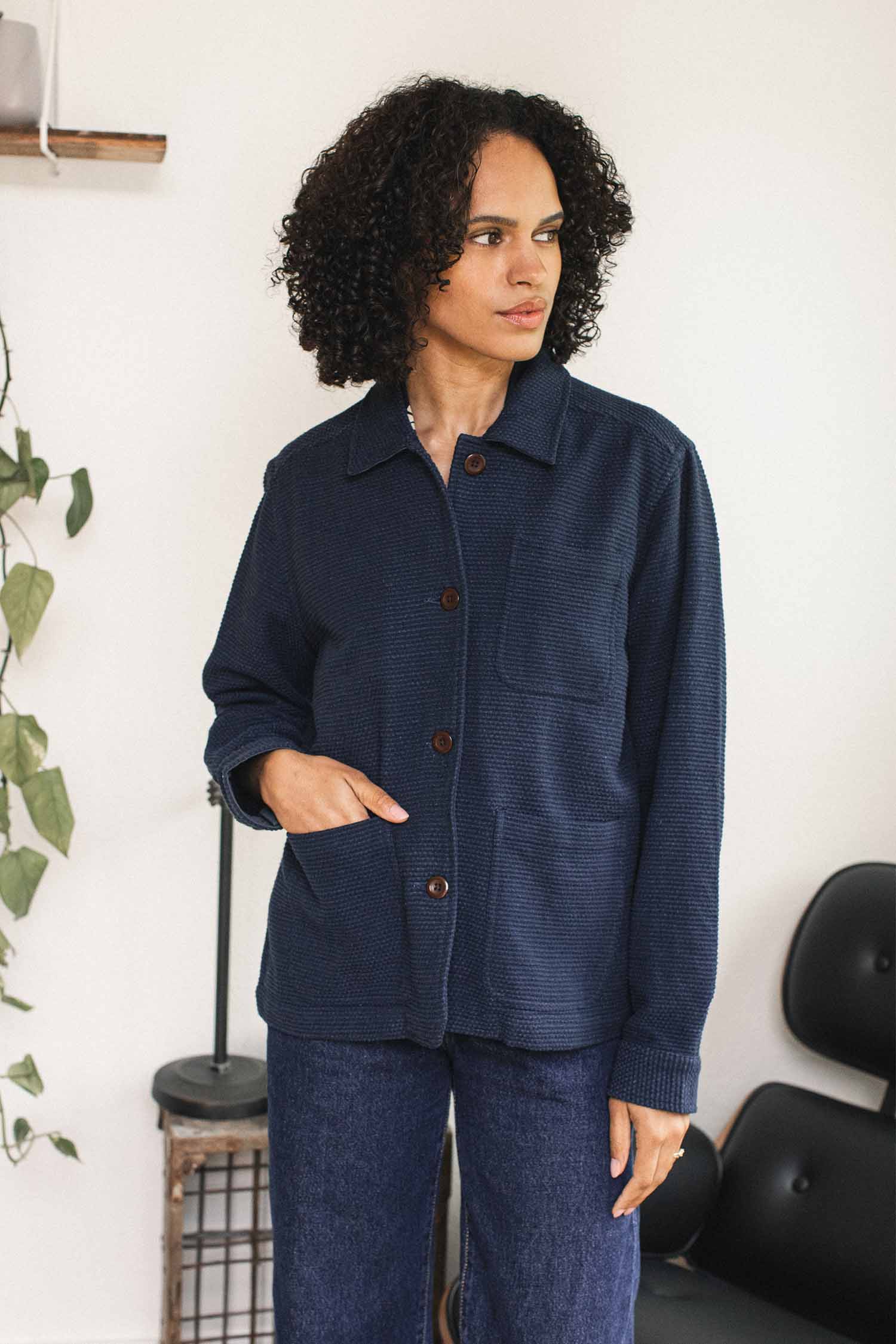 A person with curly hair wears the Bridge & Burn Women’s Studio Chore Coat in Indigo and blue jeans, standing indoors near a chair and a plant.