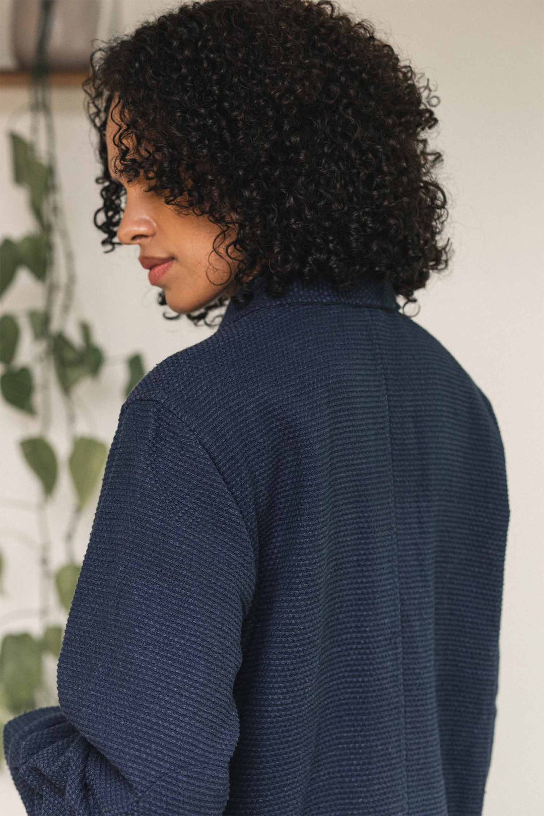A person with curly hair, wearing the Bridge & Burn Women’s Studio Chore Coat in Indigo, stands turned slightly away from the camera. A green leafy plant hangs in the blurred background.