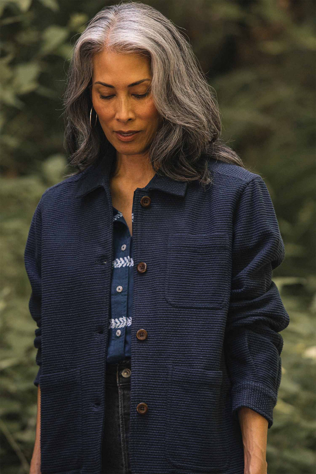 A woman with long, gray hair stands outdoors, looking down. She wears the Bridge & Burn Women’s Studio Chore Coat in Indigo over a patterned blue shirt. Green foliage is softly blurred in the background.