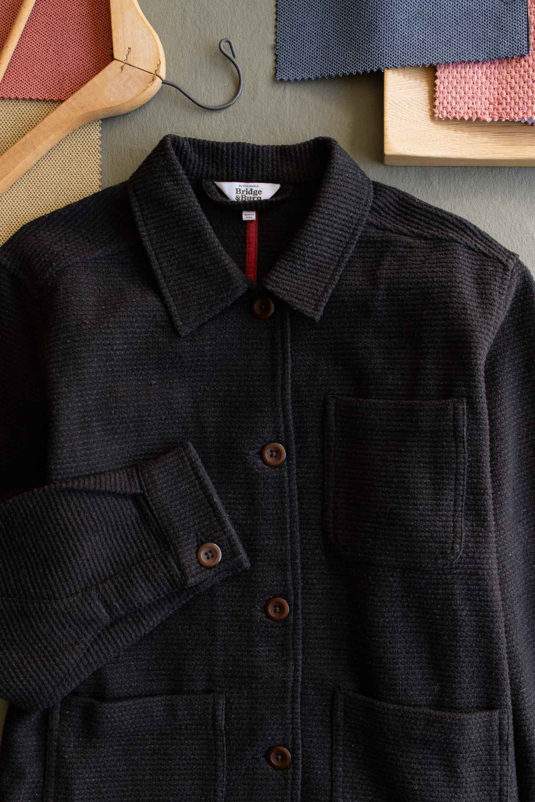 A washed black Women’s Studio Chore Coat by Bridge & Burn, crafted from organic cotton with a chest pocket, is laid flat beside fabric swatches, a wooden hanger, and a hook—perfect for workwear-inspired style.