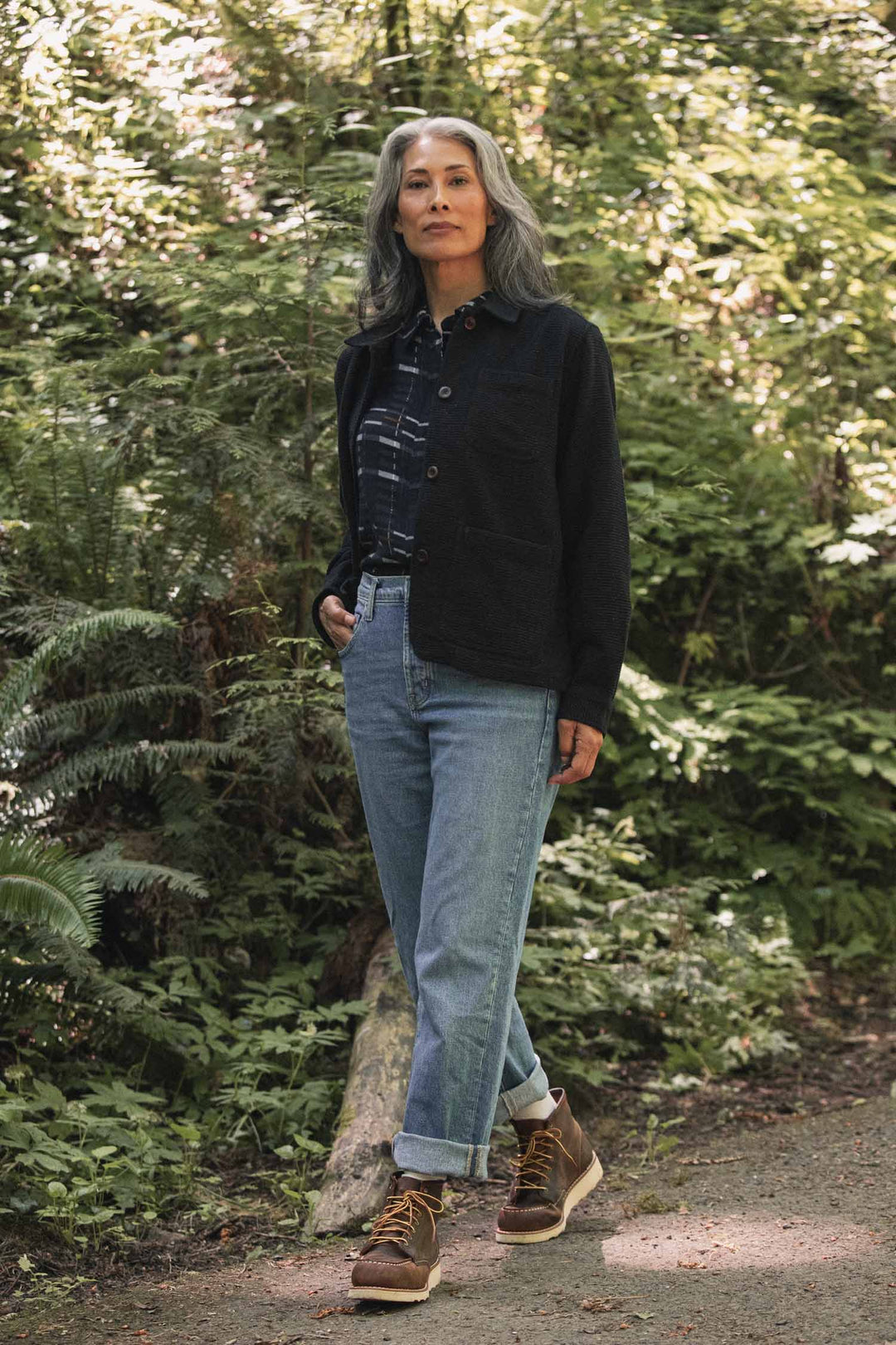 A woman with gray hair stands on a forest path, wearing the Bridge & Burn Women’s Studio Chore Coat in Washed Black over a plaid shirt, paired with cuffed jeans and brown boots, surrounded by green foliage and sunlight filtering through the trees.