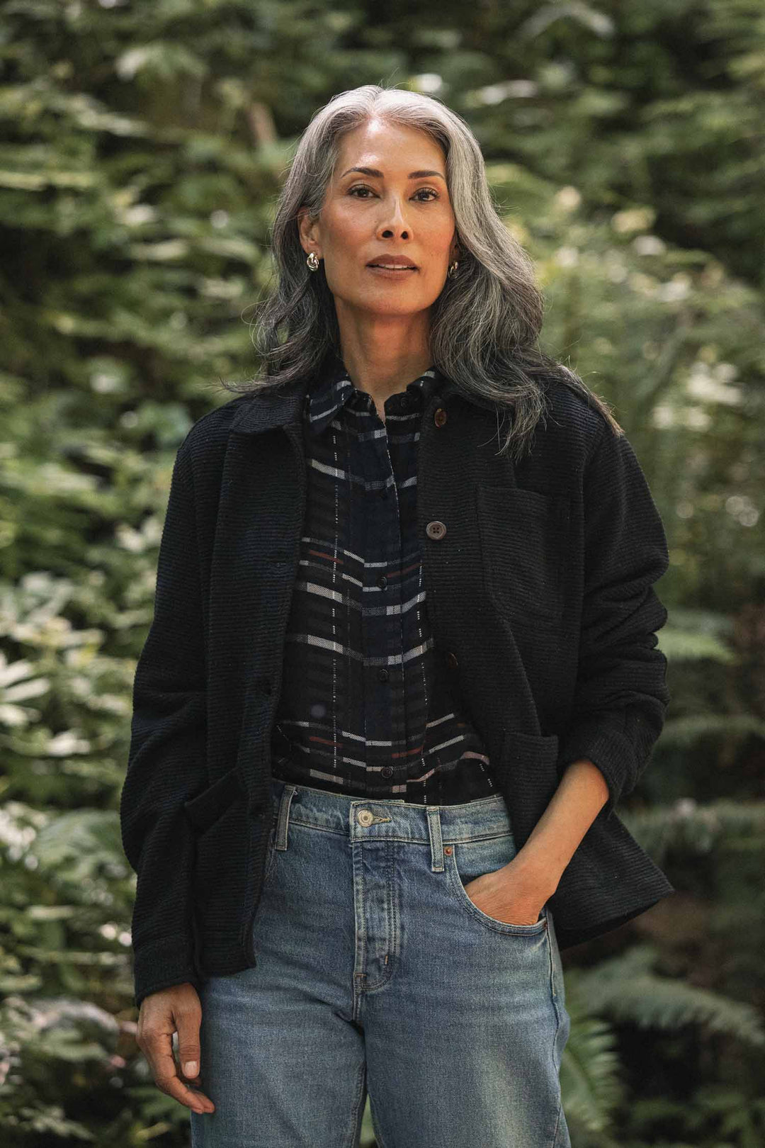 A person with long gray hair stands outdoors in a forest, wearing the Bridge & Burn Women’s Studio Chore Coat in Washed Black over a plaid shirt and jeans, one hand in their pocket, looking confidently at the camera.
