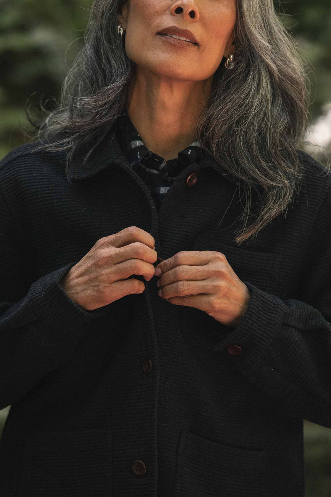 A woman with long gray hair buttons the Bridge & Burn Women’s Studio Chore Coat in Washed Black while standing outdoors. Her face is partially visible, and she wears a plaid shirt made from organic cotton underneath the coat.