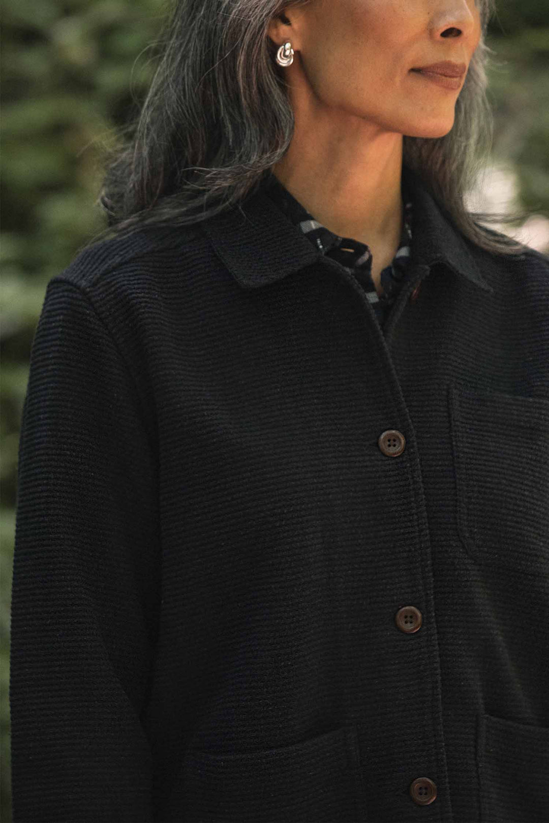 A person with long gray hair, wearing the Bridge & Burn Women’s Studio Chore Coat in Washed Black and silver hoop earrings, stands outdoors, partially out of frame.