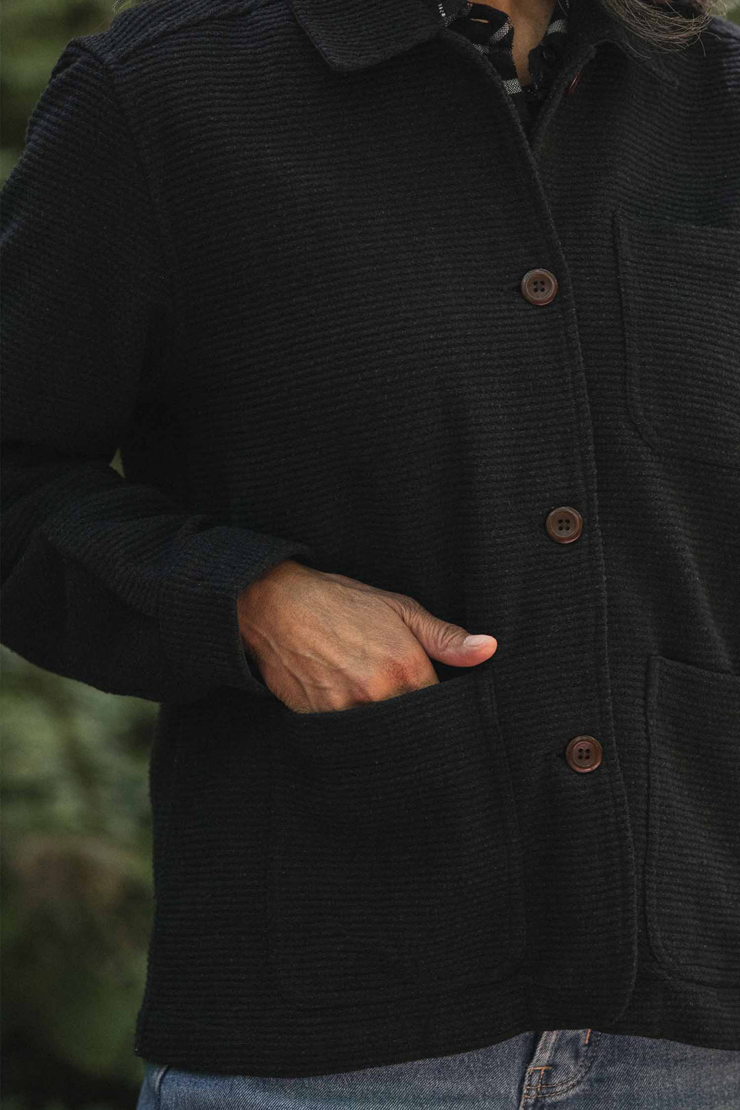 A person wearing the Bridge & Burn Women’s Studio Chore Coat in Washed Black, featuring brown buttons and a textured finish, stands outdoors with one hand in a pocket, paired with blue jeans.