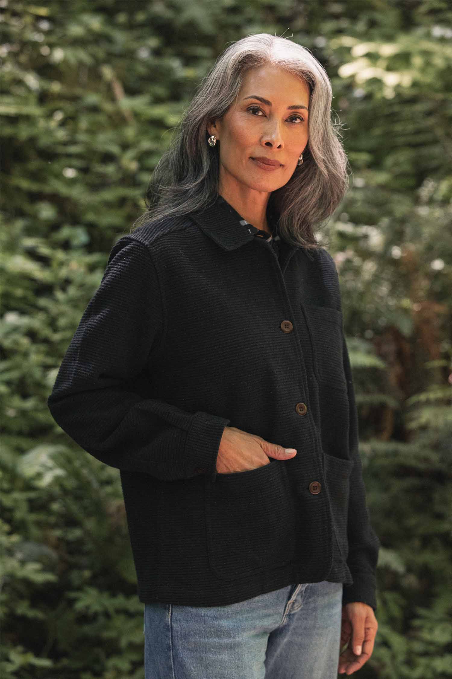 A woman with long gray hair, wearing the Bridge & Burn Women’s Studio Chore Coat in Washed Black and blue jeans, stands outdoors in front of green foliage, looking calmly at the camera with one hand in her pocket.