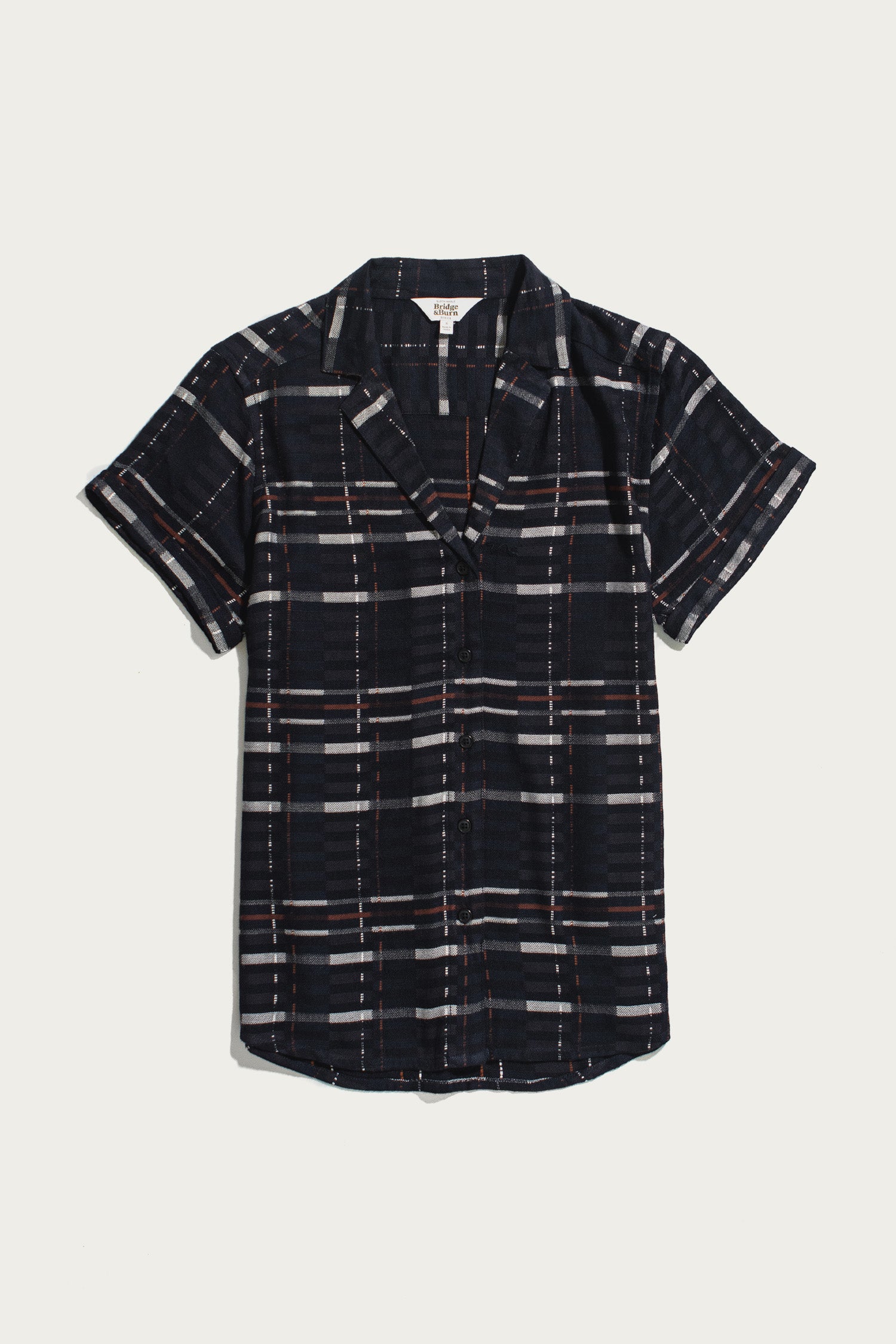 The Bridge & Burn Innes Shirt in Nightfall Stripe is a short-sleeve, relaxed fit button-up crafted from eco-friendly viscose, featuring a dark blue base with white, gray, and red plaid pattern, displayed laid flat on a white background.