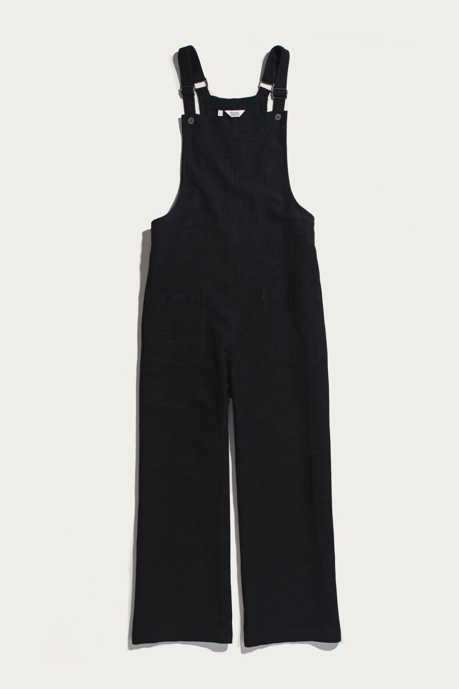 The Edie Overall in Black by Bridge & Burn is displayed flat on a white background, showcasing its wide-leg corduroy construction and relaxed fit, with adjustable shoulder straps, metal fasteners, and front pockets.
