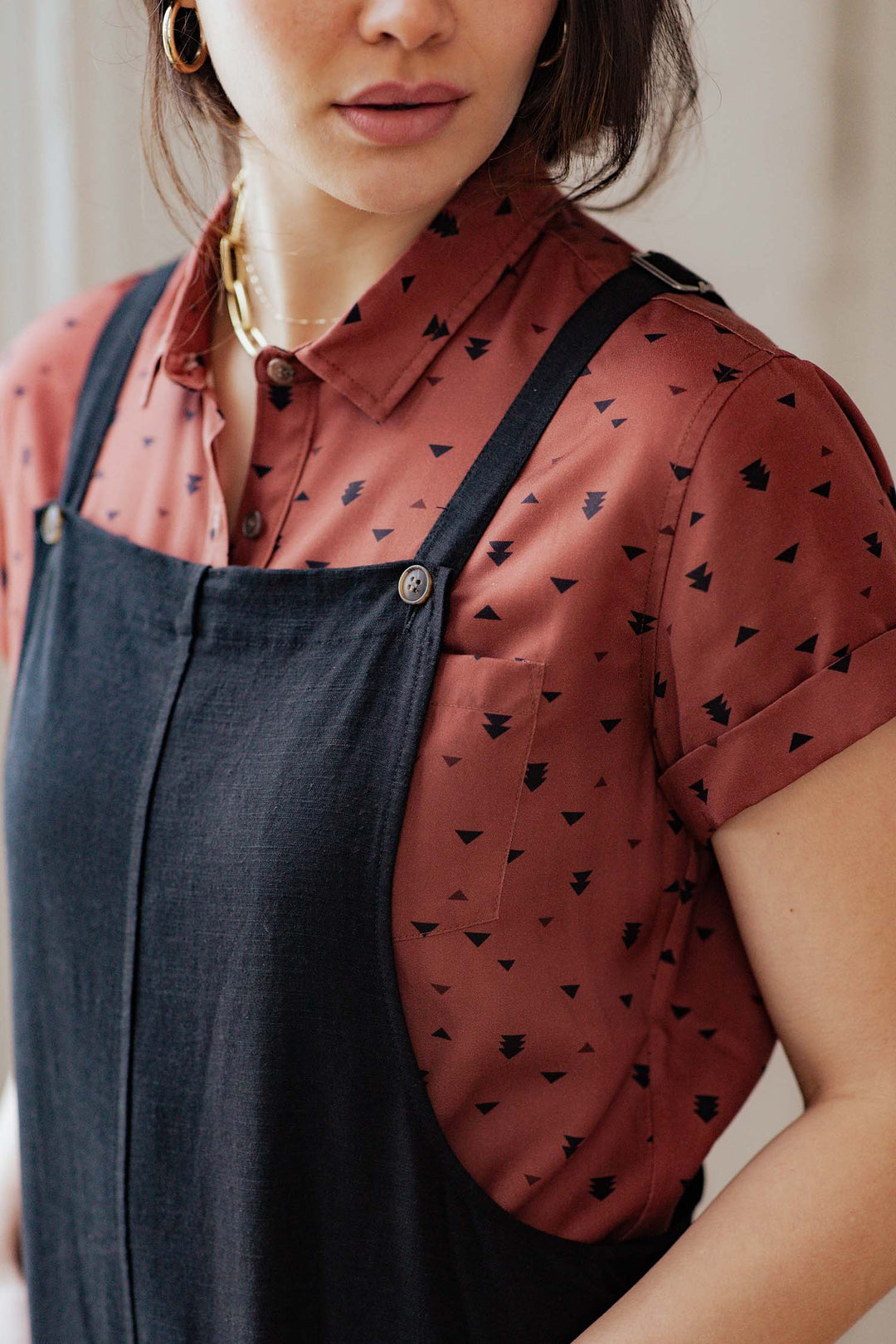 A woman wears Bridge & Burn's Edie Overall in black, styled over a rust-colored, short-sleeve button-up shirt featuring a black geometric pattern; the relaxed-fit overalls have adjustable straps and are shown from shoulder to mid-torso.
