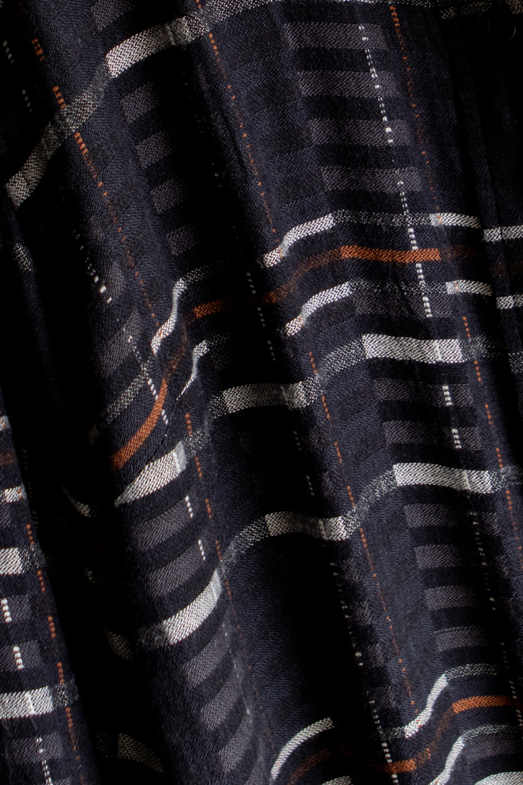 Close-up of the Innes Shirt in Nightfall Stripe by Bridge & Burn, showcasing dark eco-friendly viscose fabric with horizontal and vertical stripes in white, gray, and orange. The slightly wrinkled texture creates subtle shadows across the material.