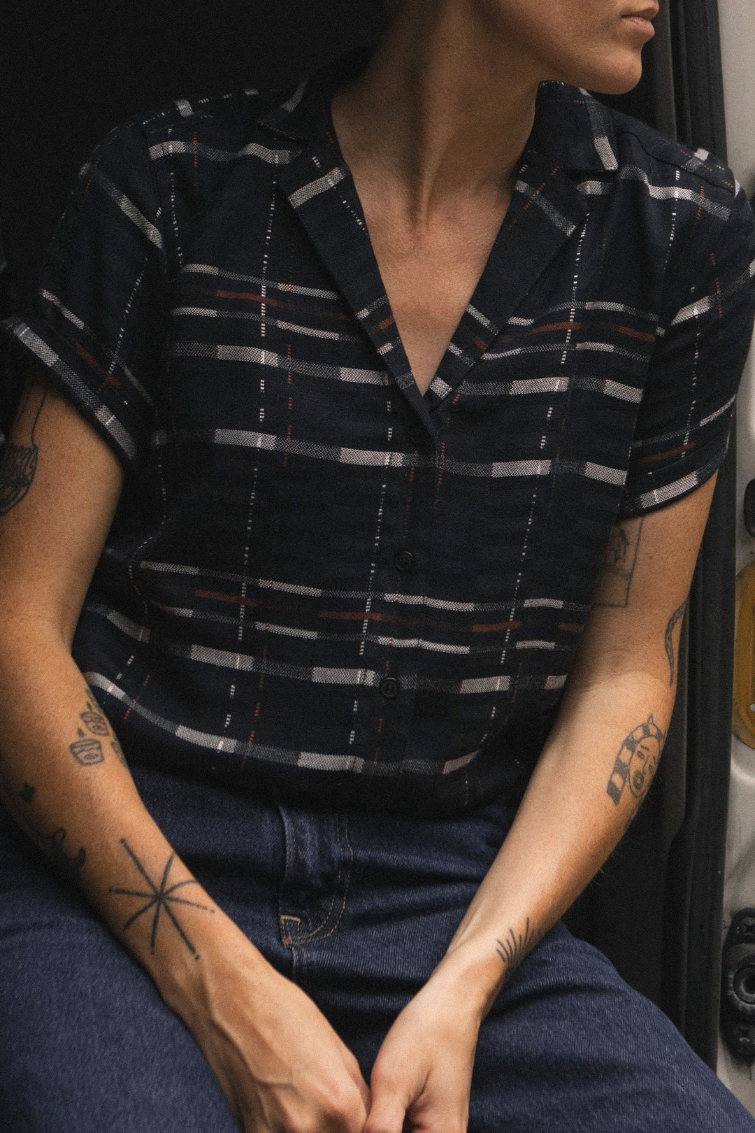 A person with short hair and visible arm tattoos wears the Bridge & Burn Innes Shirt in Nightfall Stripe—a relaxed fit, eco-friendly viscose button-up with a dark geometric pattern—paired with dark jeans. Seated with hands resting in their lap, their face is mostly out of frame.