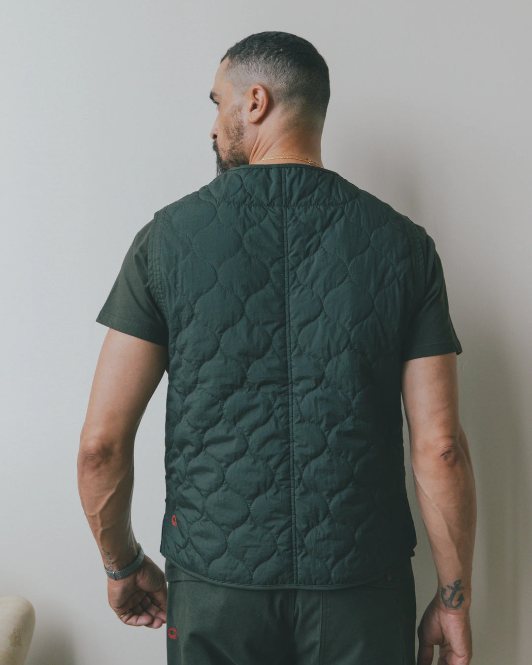 A person seen from the back wearing a dark green Imperfects - Shokunin Vest in Quilted Nylon / Greenroom over a dark green t-shirt.