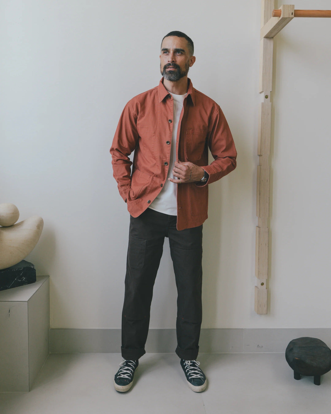 A man wearing an unbuttoned rust-colored Imperfects - Cunningham Coat in Craft Canvas / Oxide over a white t-shirt and dark trousers.