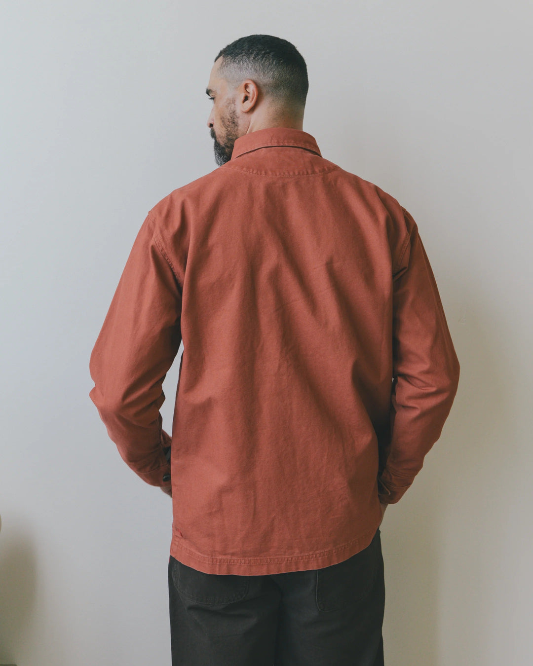 A man seen from behind wearing an Imperfects Cunningham Coat in Craft Canvas / Oxide, a long-sleeved, collared chore coat in a terracotta color.
